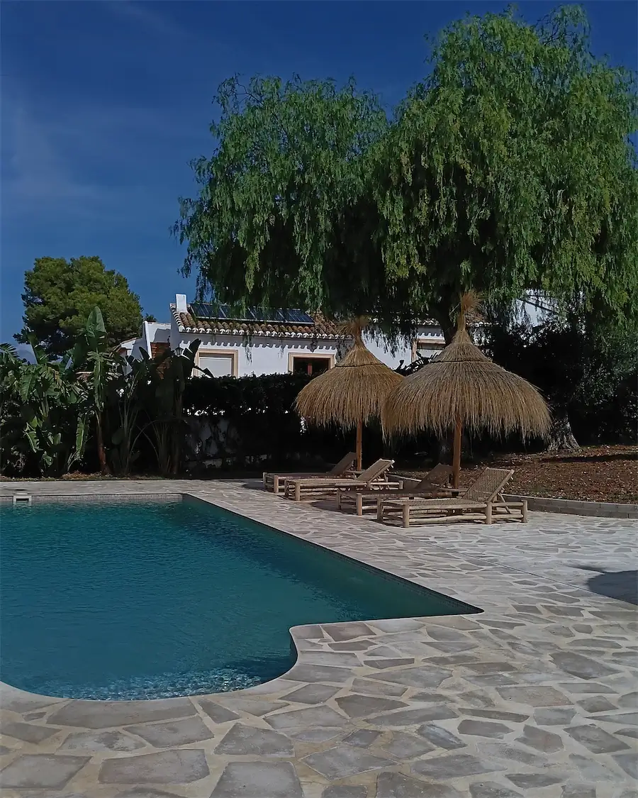 La misma piscina en octubre, con suelo de piedra, sombrillas de brezo y el árbol grande de la finca